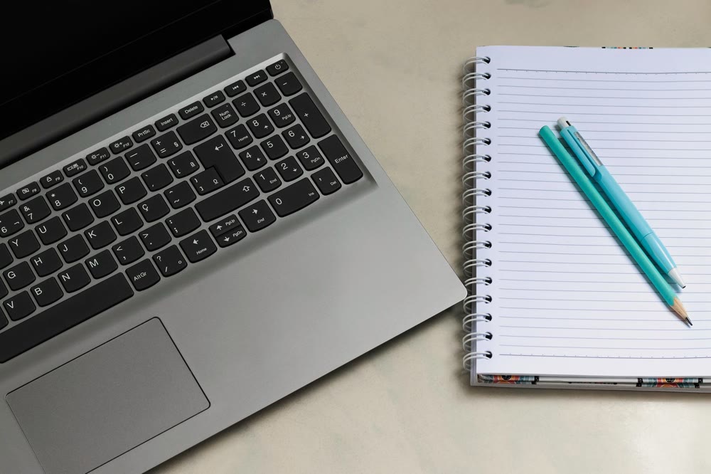 ノートPCのキーボード横に罫線入りの手帳が開かれ、水色のペンと鉛筆が置かれている机の上