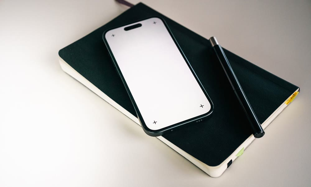 白っぽい机の上で、黒い手帳の上にスマートフォンとペンが並べて置かれている様子