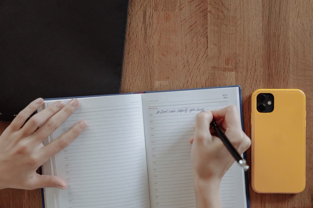 日付と時刻の欄がある手帳に書き込み、横にスマホを置いた木の机の上