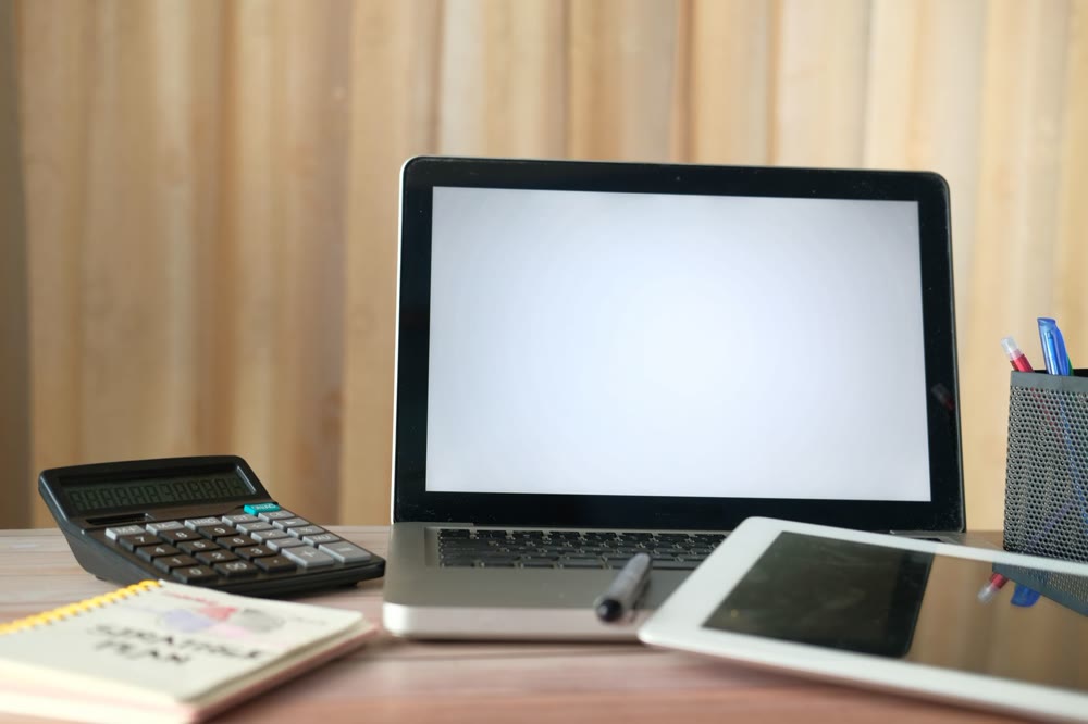 白い画面のノートパソコンと電卓、タブレット、メモ帳が並ぶ机の上