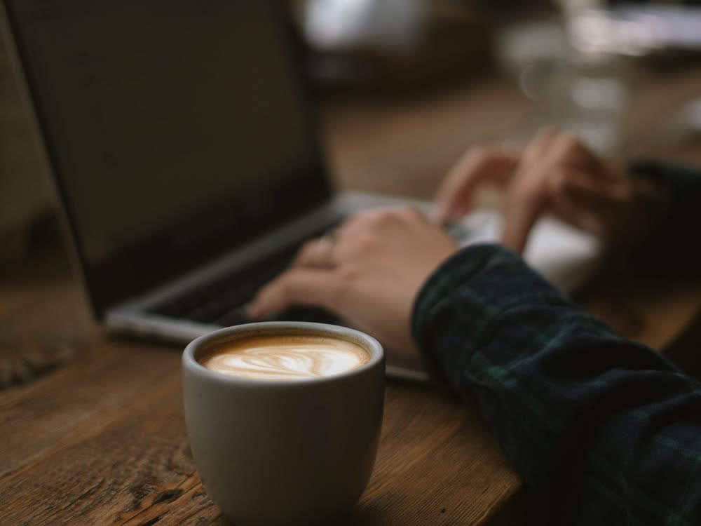 カフェのテーブルで、ラテのカップを脇にノートパソコンを打っている手元