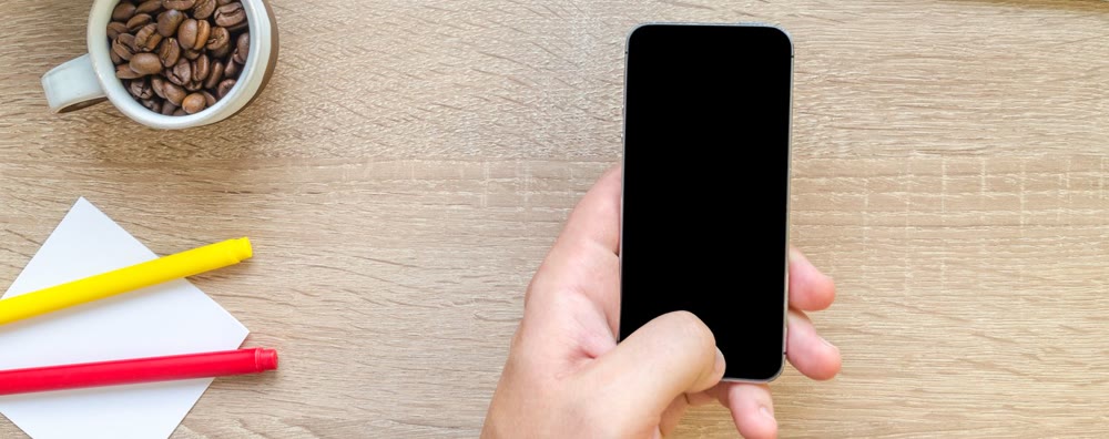 机の上でスマートフォンを片手に持つ手元と、コーヒー豆の入ったカップ、付箋、ペン
