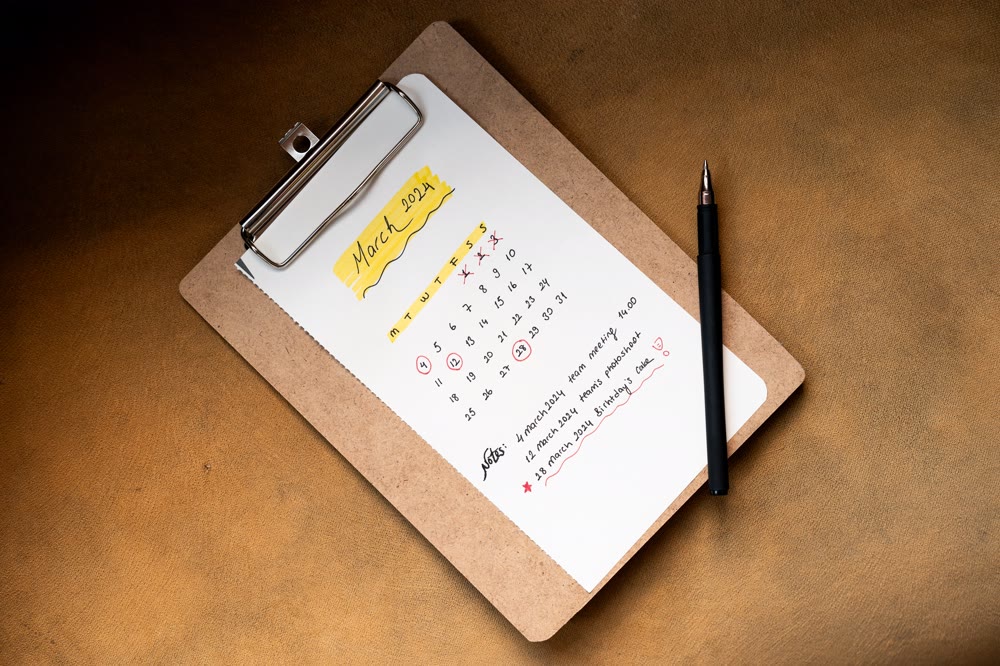 クリップボードに留めた紙に、月間カレンダーと予定が手書きされ、横にペンが置かれている様子