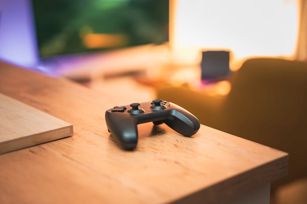 木の机の上に置かれたゲーム機のコントローラーと、奥にぼんやり見える画面