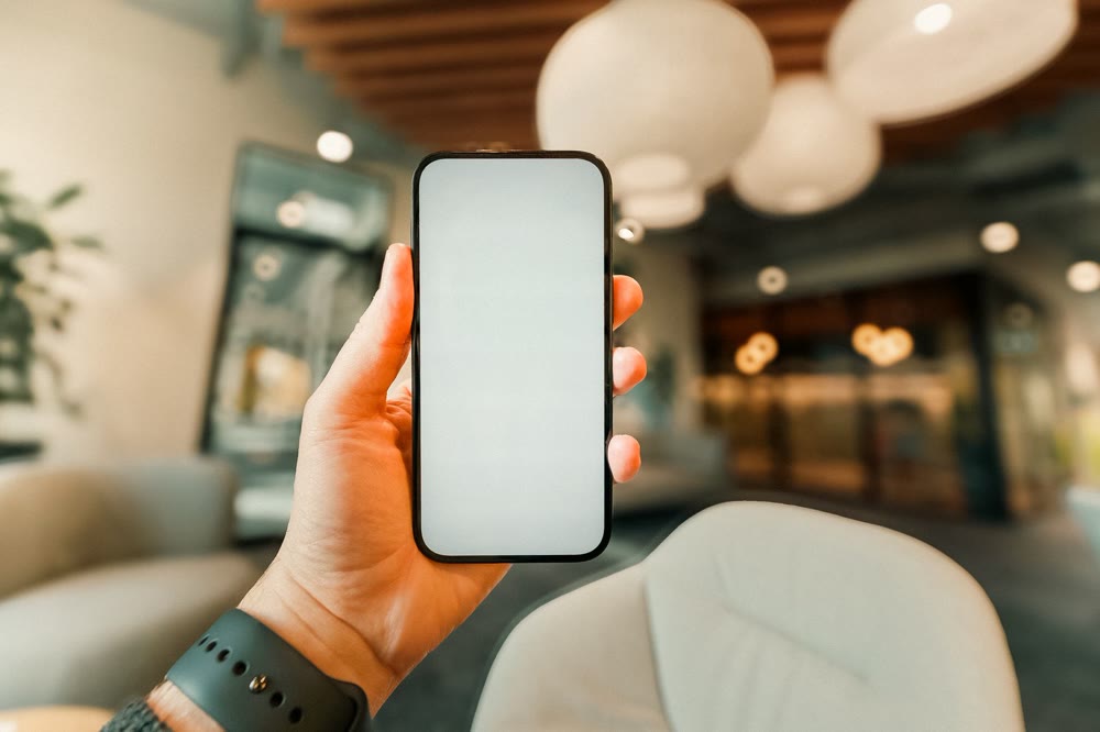 画面が無地のスマートフォンを片手に持っている、ロビーのような室内での手元