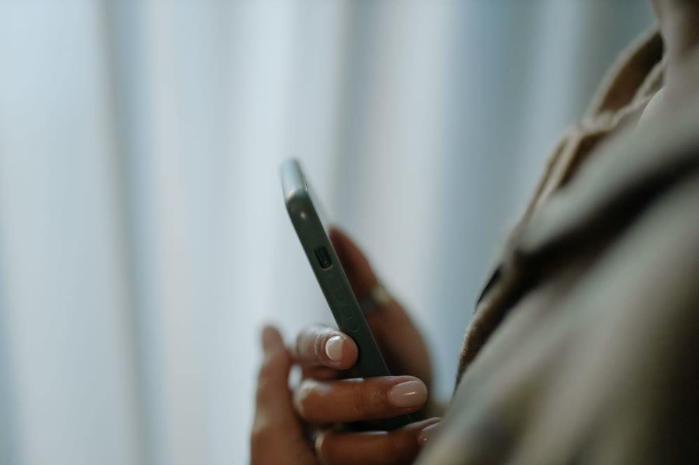 カーテンを背景に、スマートフォンを横から持って指を添えている手元