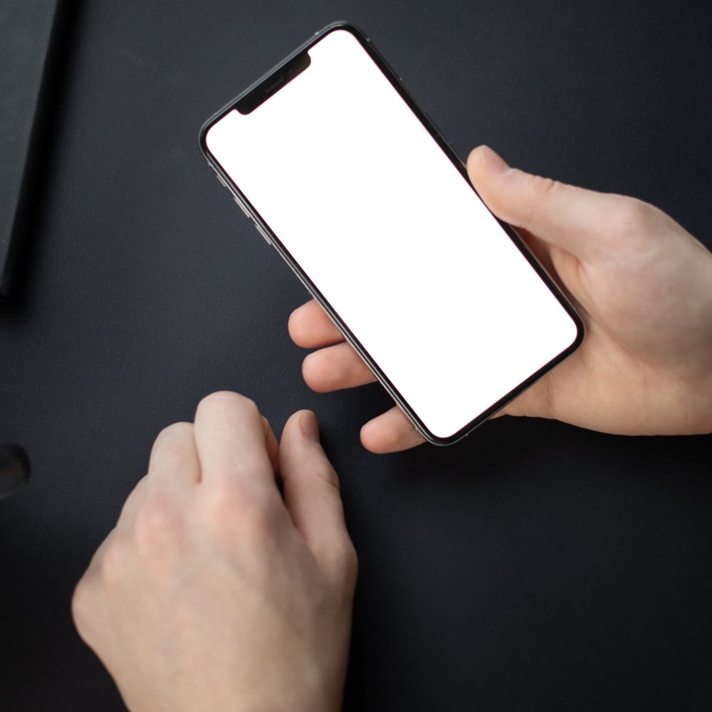 暗い色の机の上で、白い画面のスマートフォンを両手で持っている手元