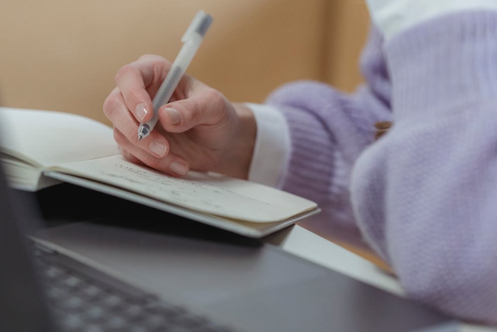 机の上でノートパソコンのそばに置いたノートへ、ペンで書き込んでいる手元
