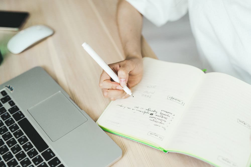 ノートパソコンの横で、ノートにペンで項目を書き出している手元